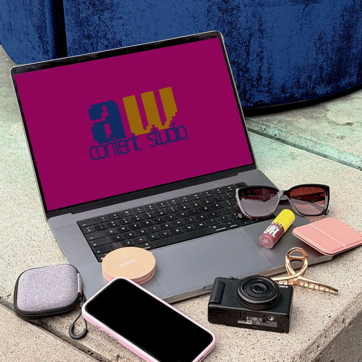 Laptop z logo AW Content Studio na magentowym ekranie, obok aparat, okulary przeciwsłoneczne i kosmetyki